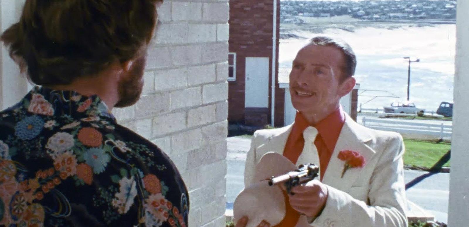 A man with his back to camera stands in a doorway to a building facing another man in a white suit who is pointing a gun at him.