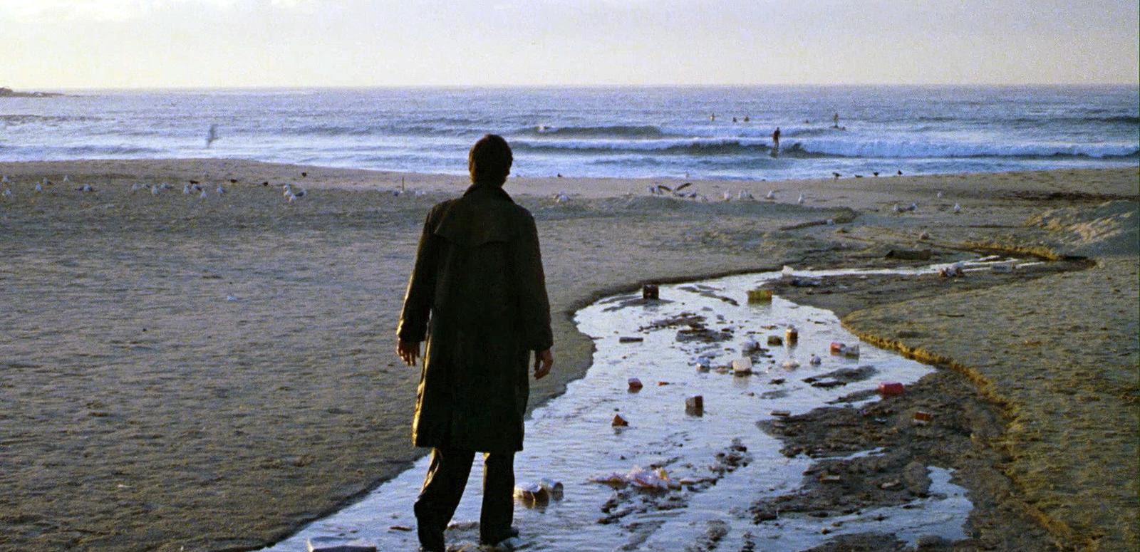 A man stands alone on an empty beach, wearing a long coat and with his back facing the camera. There is rubbish strewn on the beach in front of him.