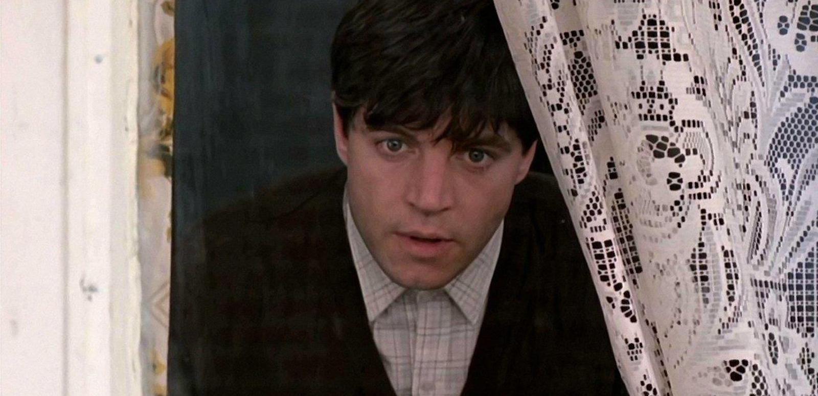 A young man peering out of a window through curtains.