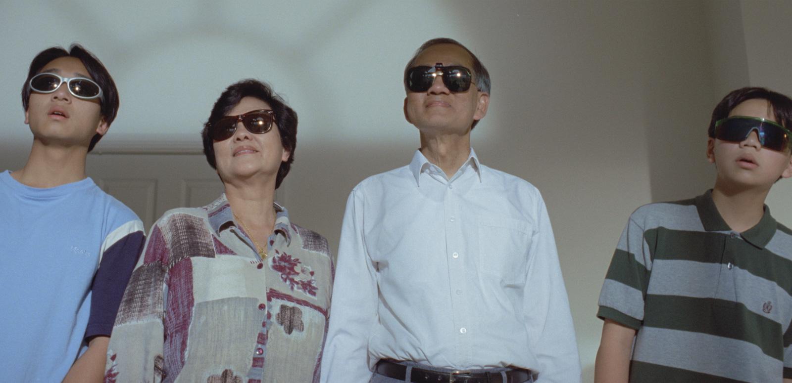 A family of four people standing side by side in a room looking outside. They are all wearing dark sunglasses.
