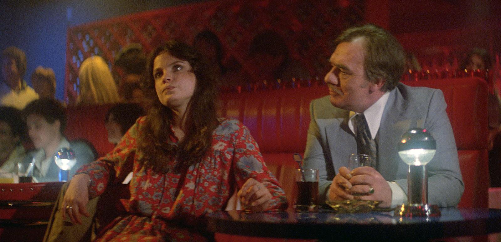 A young woman sits at what appears to be a table in a bar. She is looking unhappy with her head tilted away from an older man who is talking to her.