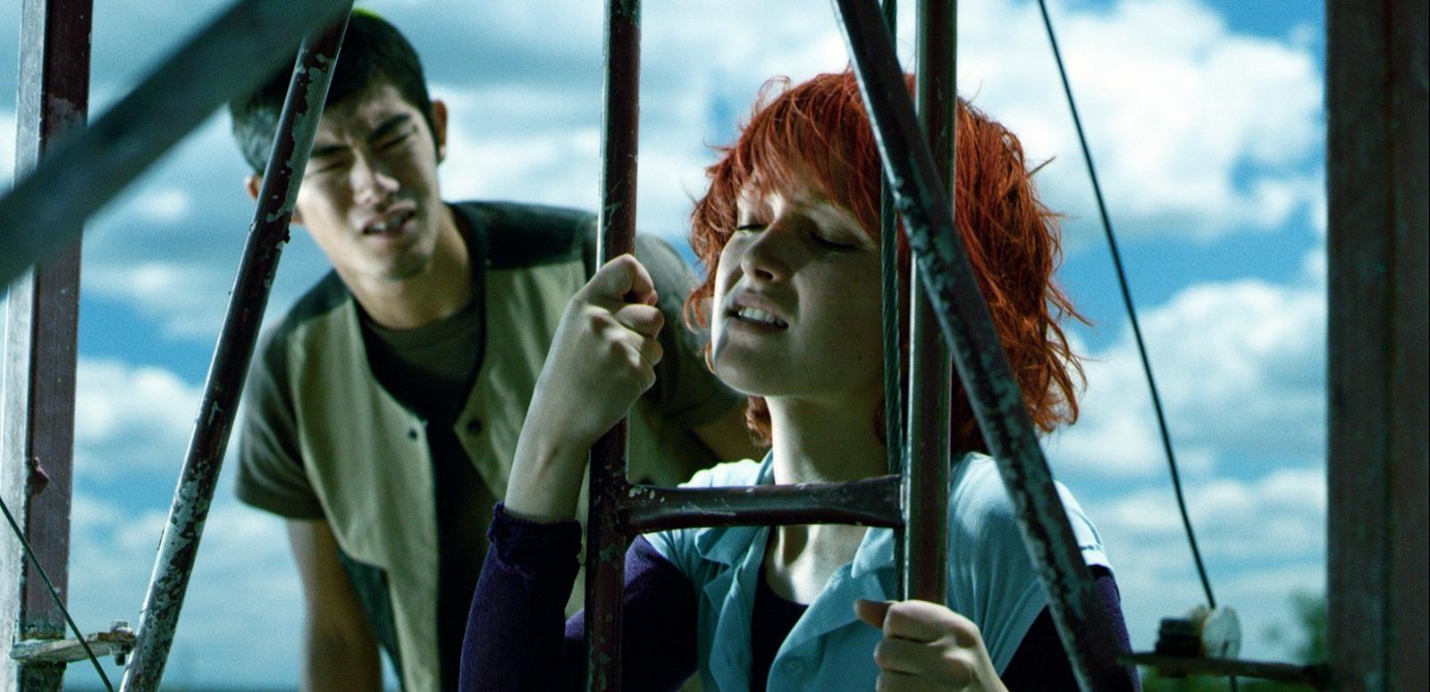 A confused-looking young man looks at a young woman who has her eyes closed and is holding on tightly to a metal ladder.