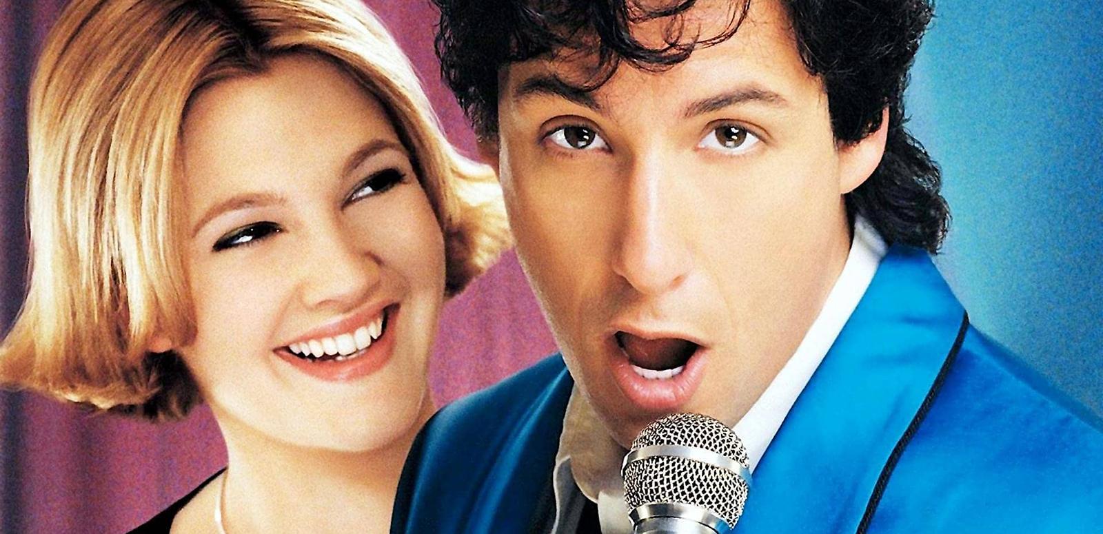 Close up of a young woman with short blonde hair (Drew Barrymore) smiling and looking at a man (Adam Sandler) singing into a microphone in a promotional shot for the film The Wedding Singer.