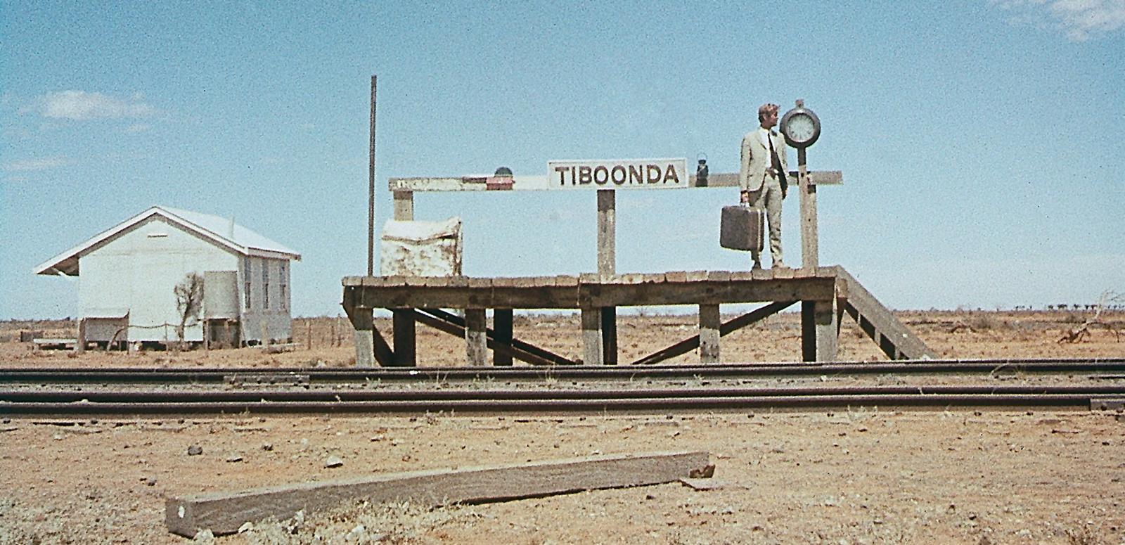 Wide shot of a man holding a suitcase is standing on a short platform at a train station in a remote outback area. The sign at the train station says Tiboonda.