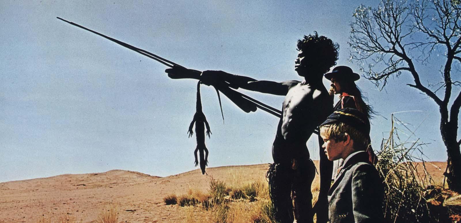David Gulpilil, pointing a spear, stands in the desert with Jenny Agutter and Lucien John in private school uniforms in a scene from the film Walkabout