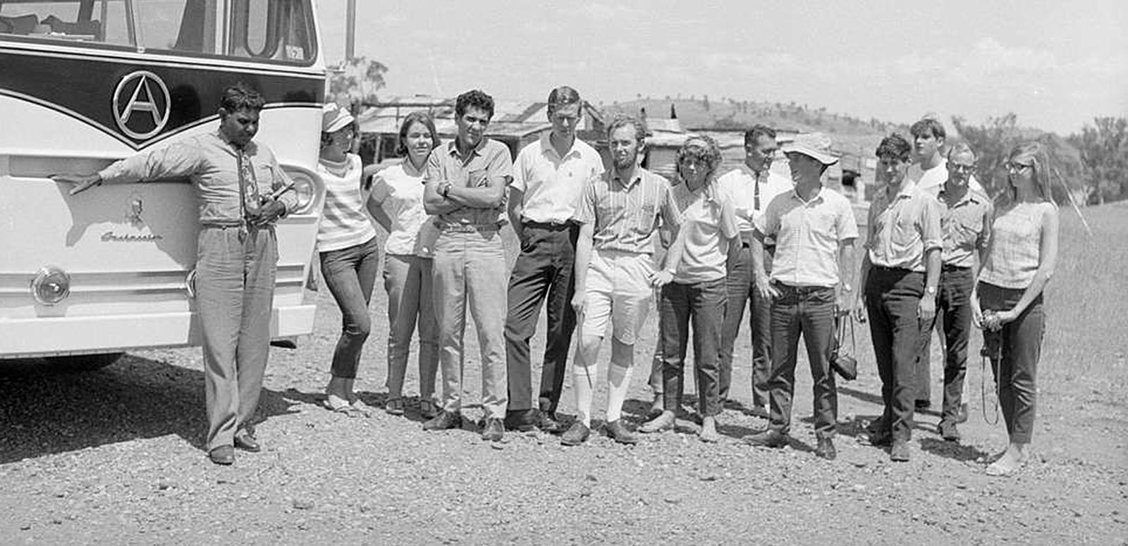 A group of 13 people standing side by side next to the front of a bus looking towards the camera.