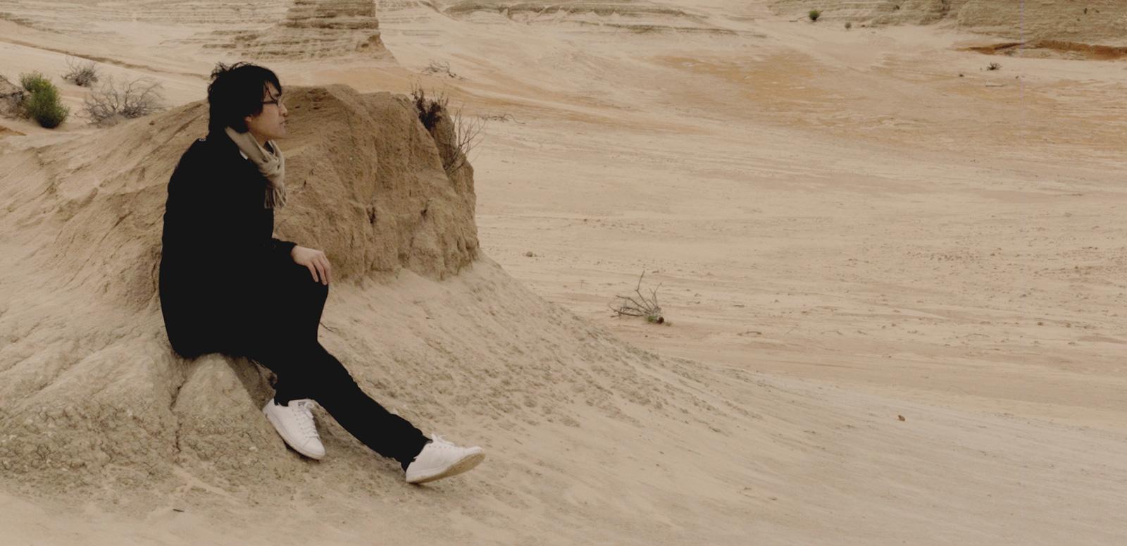A young Asian man sitting on a rock in sparse desert setting.