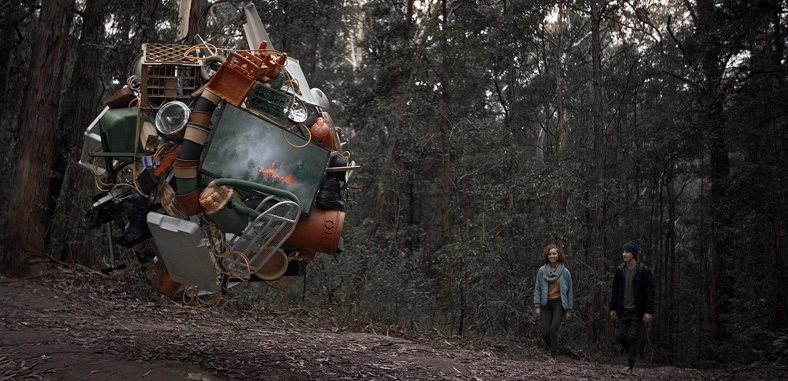 A young man and woman walking through a wooded area, in the foreground is a large spherical hanging structure made up of various items including pots, buckets, crates, bins and other different metal and plastic objects.