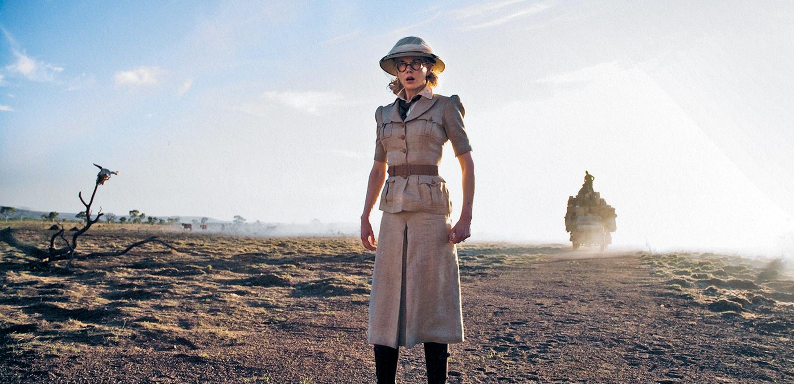 Full-length, wide shot of actor Nicole Kidman in a scene from the film Australia. She's standing in the dusty outback wearing a safari-style outfit of a beige skirt, belted shirt and hat with long dark brown boots. 