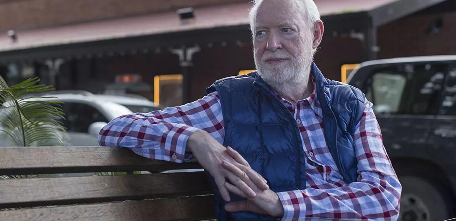 Film critic and author David Stratton seated on a bench outdoors. He is wearing a check shirt and vest and looking off to his right.