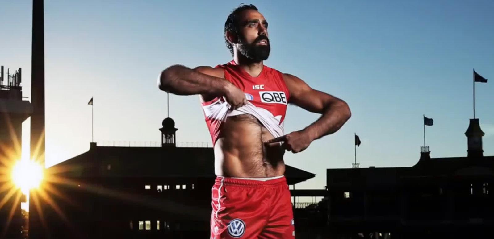 AFL football player Adam Goodes in the middle of a sports ground. He is wearing his Sydney Swans red and white uniform and lifting up the front of his shirt to point to the colour of his skin. 