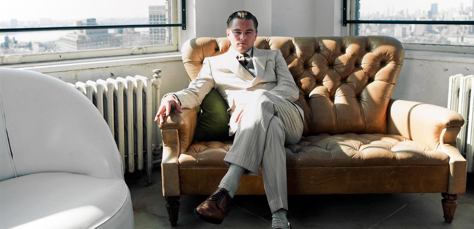 Actor Leonardo DiCaprio in a behind the scenes still from the film The Great Gatsby. He is wearing a white suit and is seated on a tan-coloured couch in the corner of a room. He's looking directly at the camera. Out of the windows behind him you can see a city skyline. 