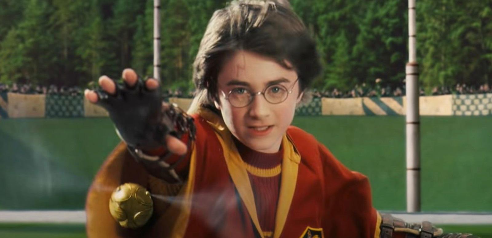 A young teenage boy wearing maroon and gold robes with his arm outstretched trying to catch a small golden ball in a scene from Harry Potter and the Philosopher's Stone.