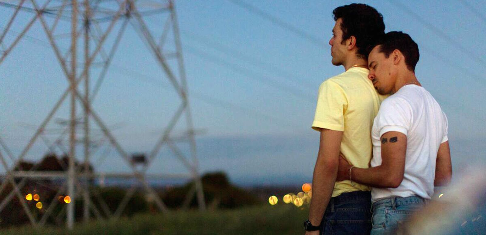 A young man holds another young man around the waist from behind. They are standing outside with a view of an electrical tower