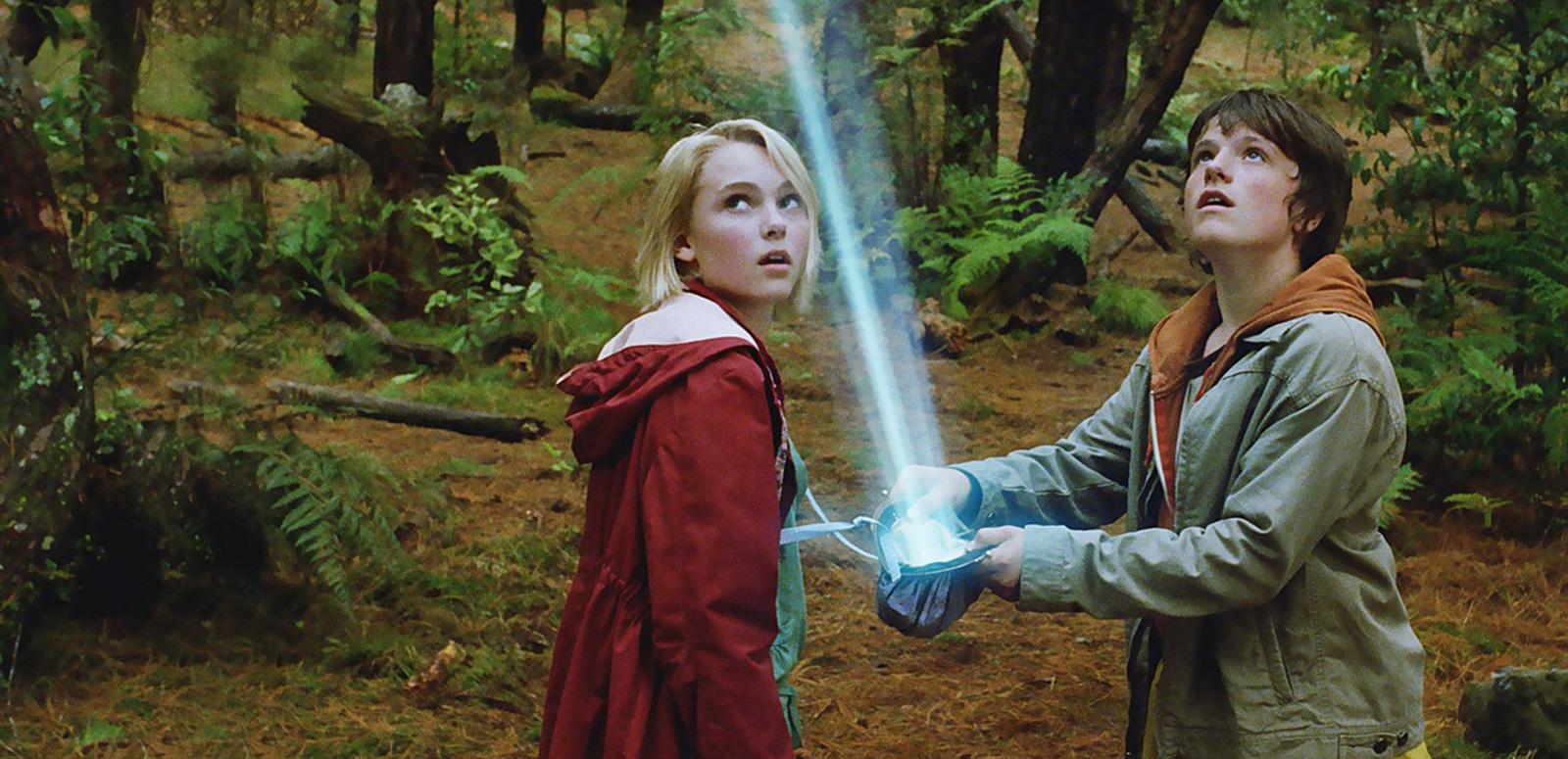 Two young teens are in a forest looking up towards the sky at a bright light beaming down in front of them.