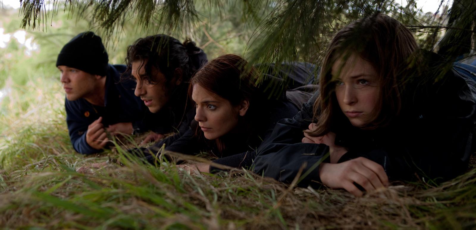 Four teenagers lying on the ground, trying to hide themselves under trees and scrub as they watch over something happening off screen.