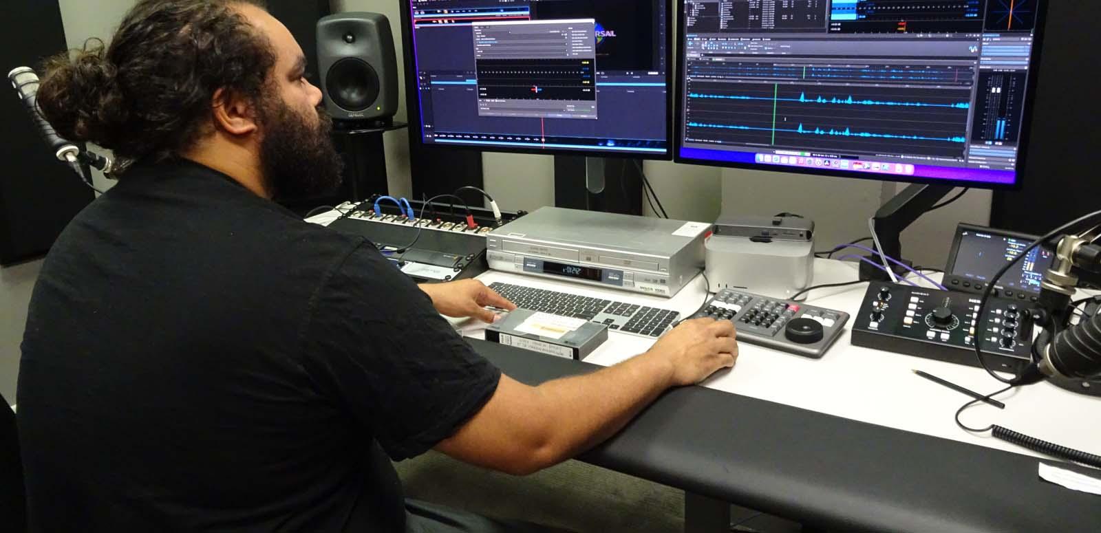 A First Nations man seated at a desk in a studio covered in computers and recording equipment