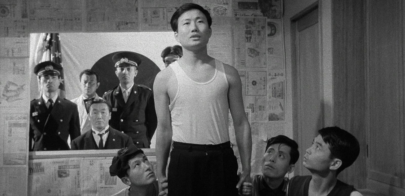 A young Japanese man standing in a room surrounded by three other men who are holding onto his hands. In the back of the room are a group of policemen.