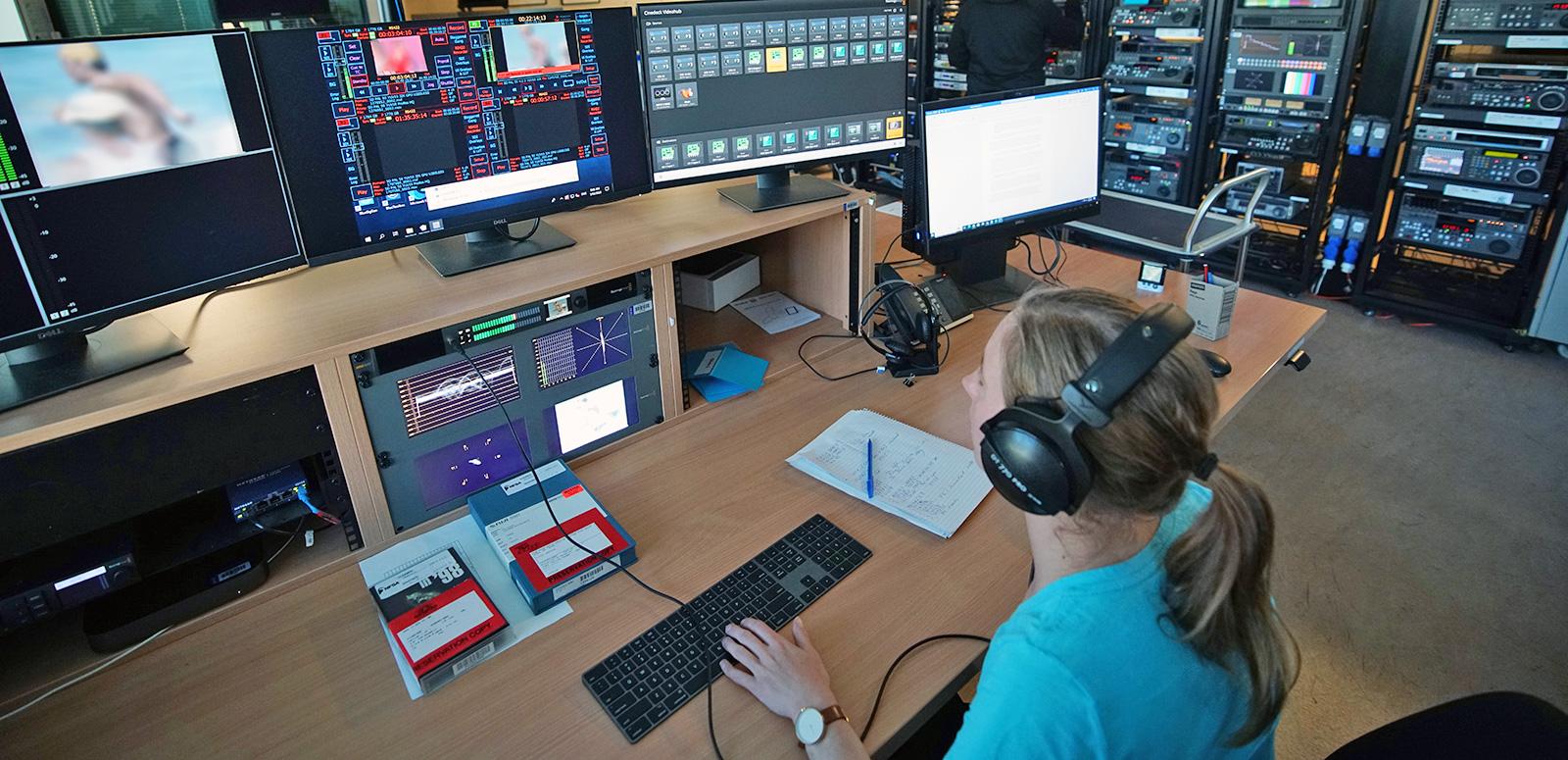 A woman sitting at a desk wearing headphones. She is looking at a number of computer monitors and working on a keyboard. 