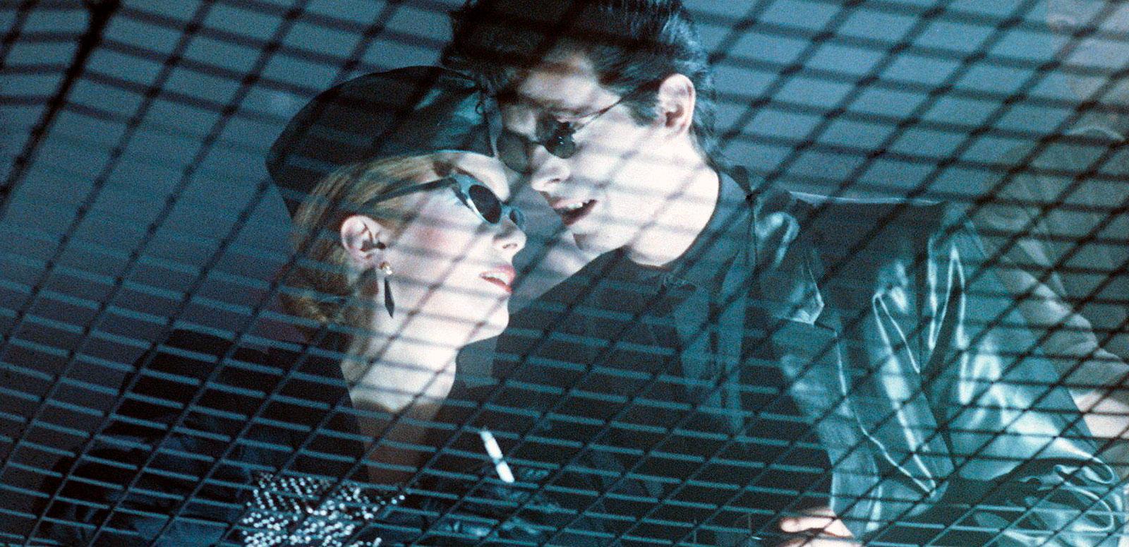 A scene from the film The Hunger featuring Catherine Deneuve and David Bowie as vampires. They are pictured looking up from below through a wide net, wearing dark clothing and sunglasses at night and standing close together.