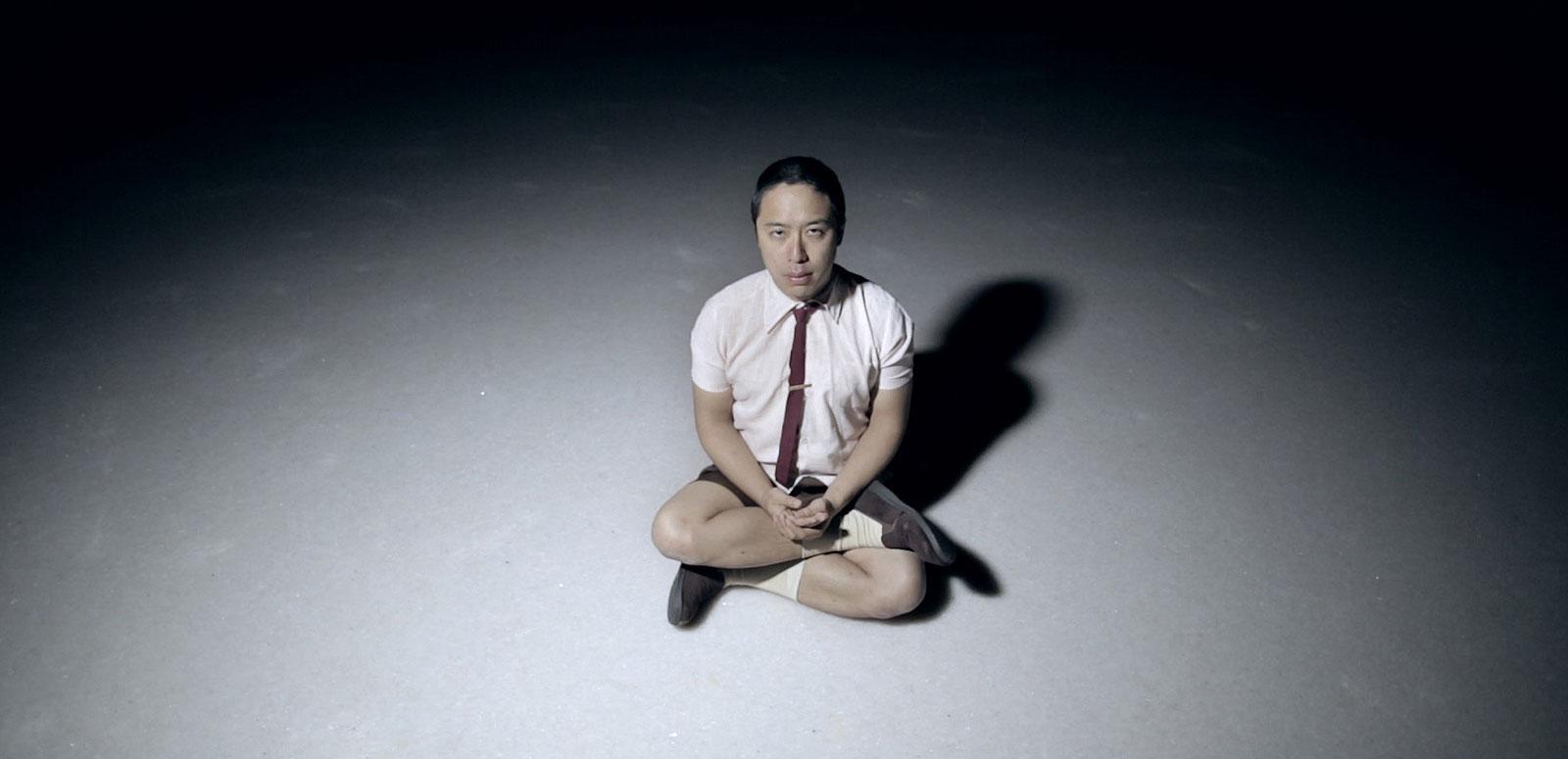An Asian man sitting cross-legged on a floor in a dark room with a spotlight on him. He is wearing a short-sleeve shirt with a tie and shorts and he is looking up at the camera.