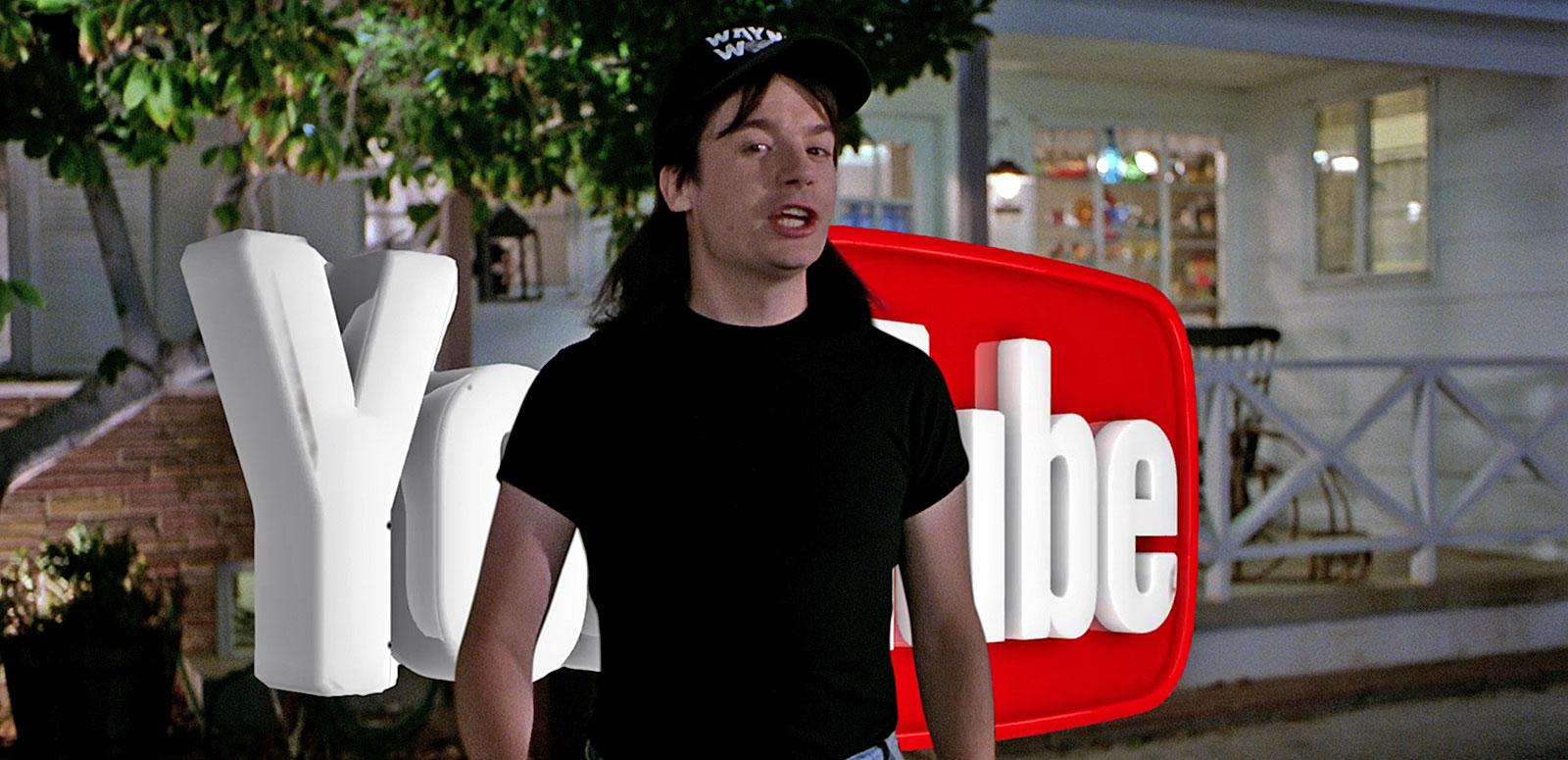 Mike Myers dressed as his character from the film Wayne's World pictured in front of a big YouTube sign.