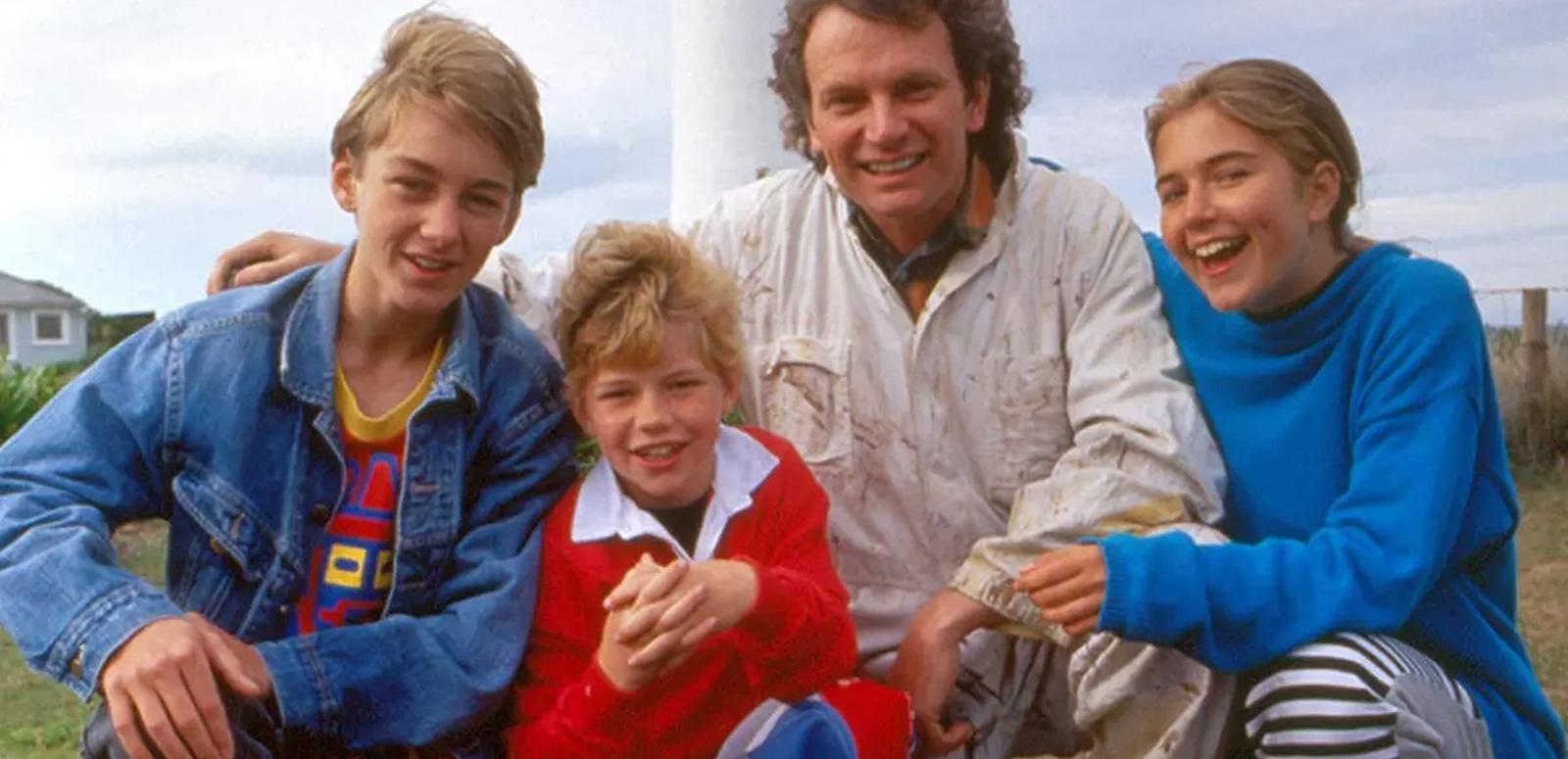 A family of four - two boys, a girl and their dad, kneeling with their arms around each other and smiling at the camera. You can see part of a lighthouse in the background. 