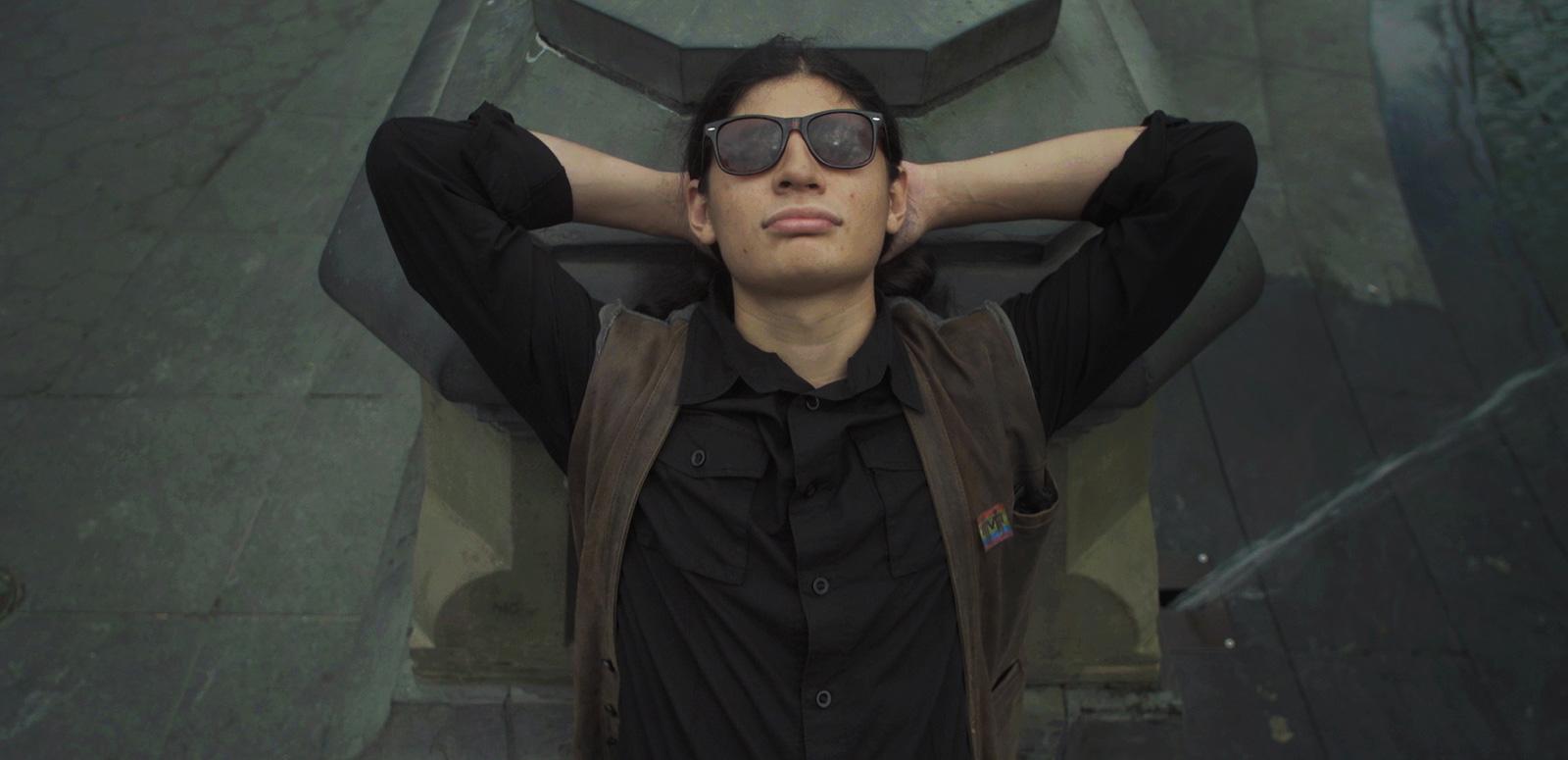 A young man pictured from the waist up wearing dark sunglasses, leaning back with his arms raised and hands folded behind his head.
