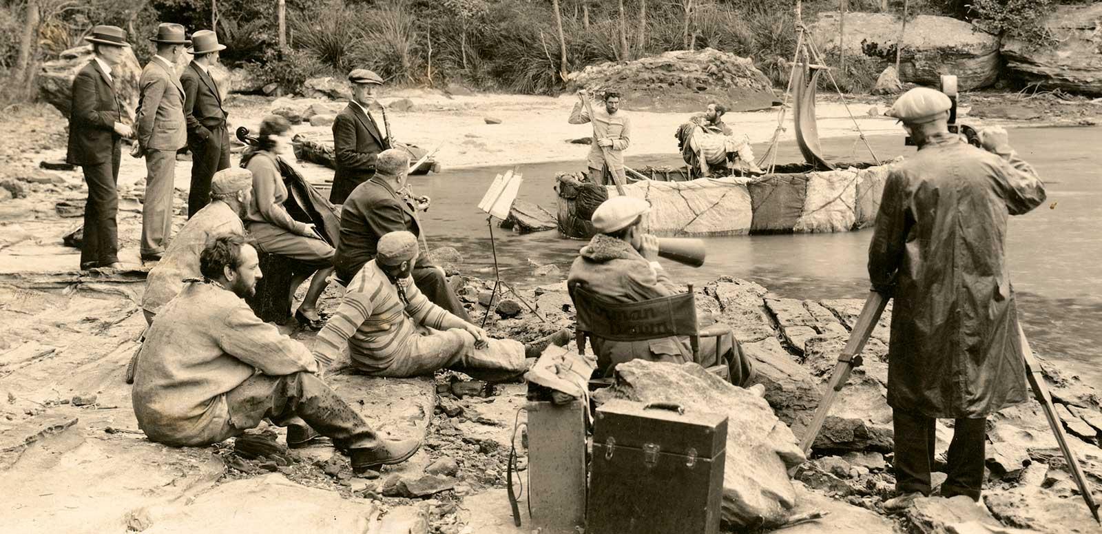 A film crew and actors on a rocky beach filming a scene in which two men dressed as convicts and a woman are on a makeshift boat.