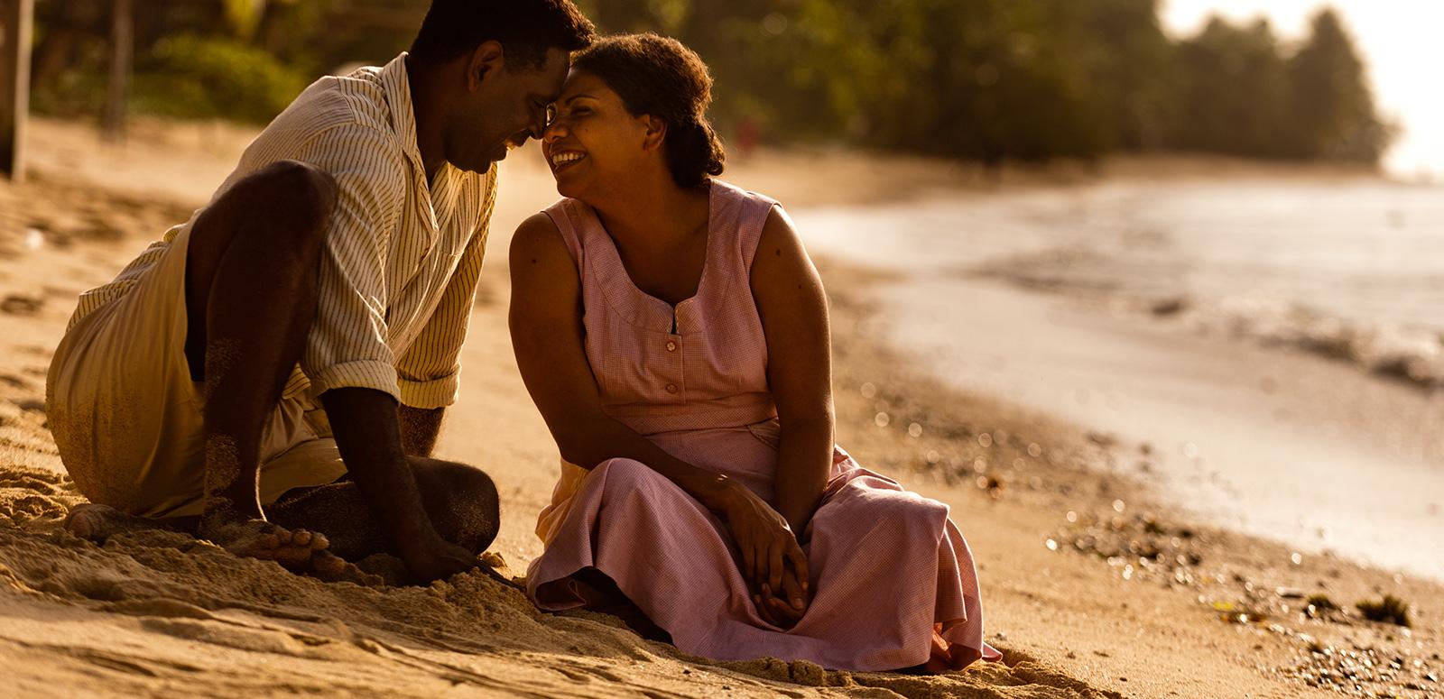 A First Nations man and woman sit together on a beach looking at each other and smiling in a scene from Mabo.