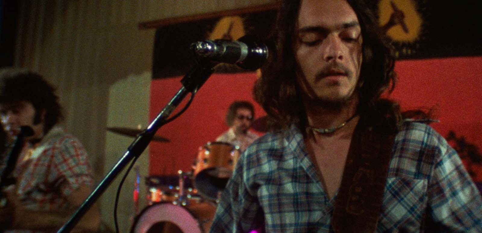A First Nations man wearing a checked shirt and playing guitar on stage with a rock band in front of an Aboriginal flag.