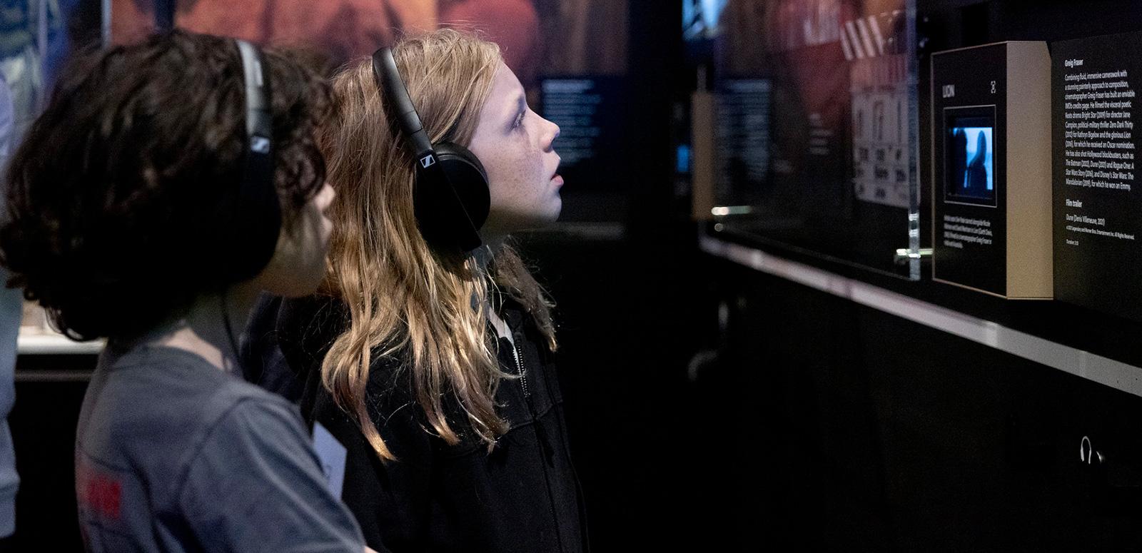 Two kids wearing headphones and looking at writing and a screen on the wall of an exhibition.