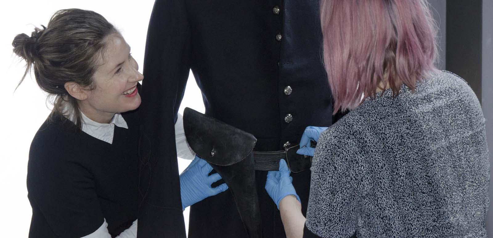 Two curators at the NFSA adjust the belt on a costume on a mannequin. One of the curators is a young woman who is laughing while she works, and both are wearing blue gloves.