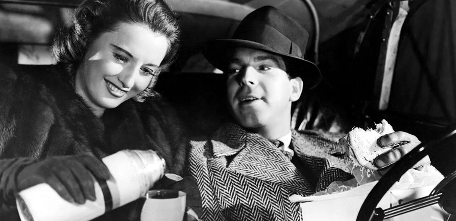 A woman and man sitting side by side. They are both wearing warm coats and gloves. The woman is pouring a drink from a thermos into a cup and smiling and the man is looking at her while holding a sandwich in one hand.