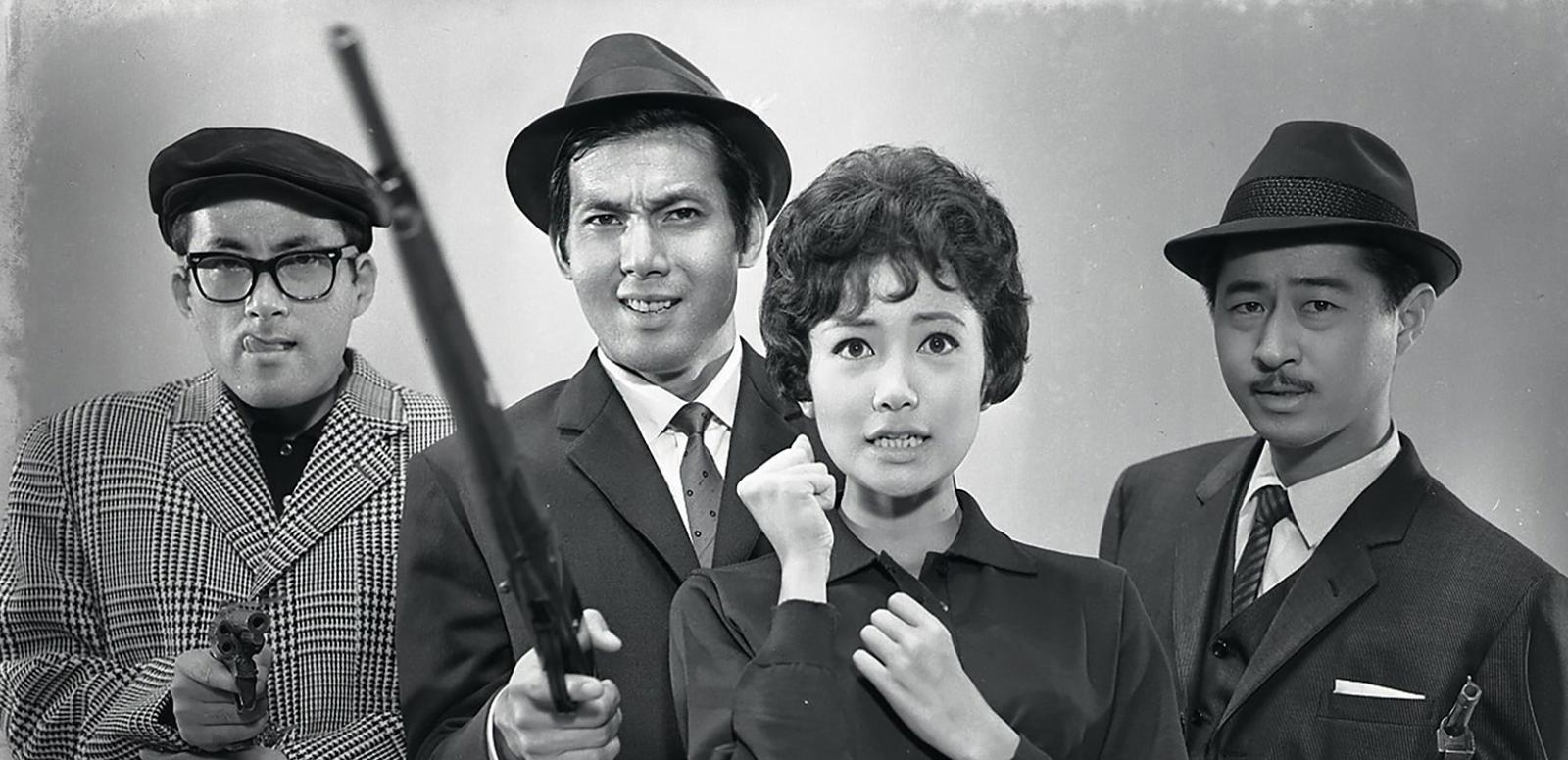 Four Japanese people - 3 men and a woman - pictured standing side by side looking towards the camera. The men are all wearing suits and hats and holding firearms.