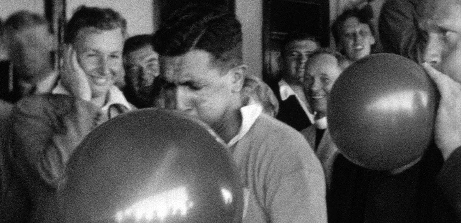 Two men blowing up balloons while a group of other men standing in the background are watching