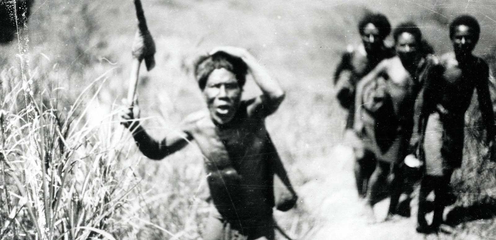 A group of four Indigenous men walking through a grassy area. One of them is holding a tool of some description in one hand, with his other hand on his head.
