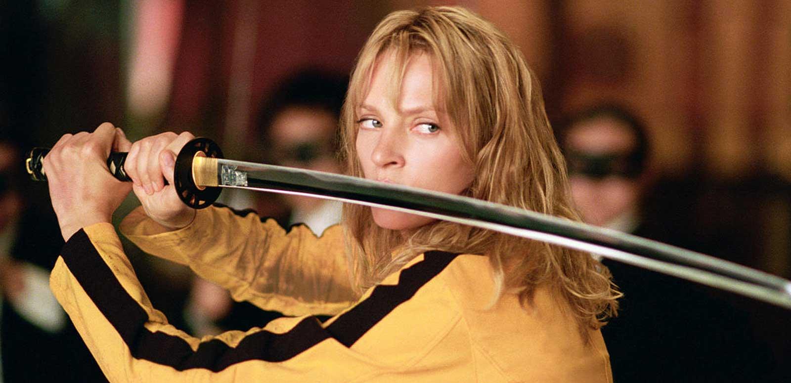 Close up of actor Uma Thurman holding a sword in both hands and wearing a yellow tracksuit with a black stripe in a scene from Kill Bill Volume 1.