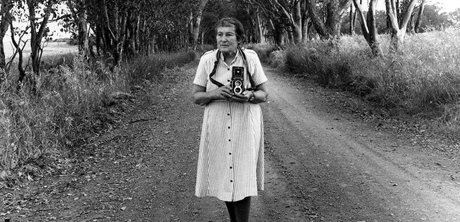 Wide shot of an older woman standing in the middle of a dirt road that is lined by trees. She is holding an old camera.