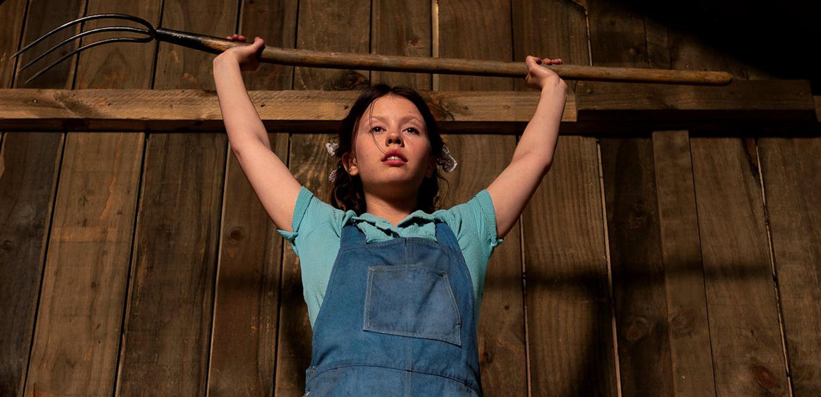 An angry-looking teenage girl standing in what looks to be a barn, is holding a pitchfork above her head.