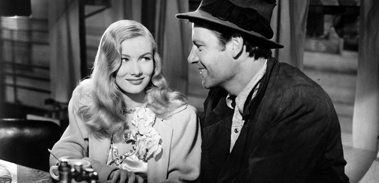 Actors Veronica Lake and Joel McCrea sitting next to each other in what appears to be a bar or a diner. They are smiling at each other and leaning in towards each other.