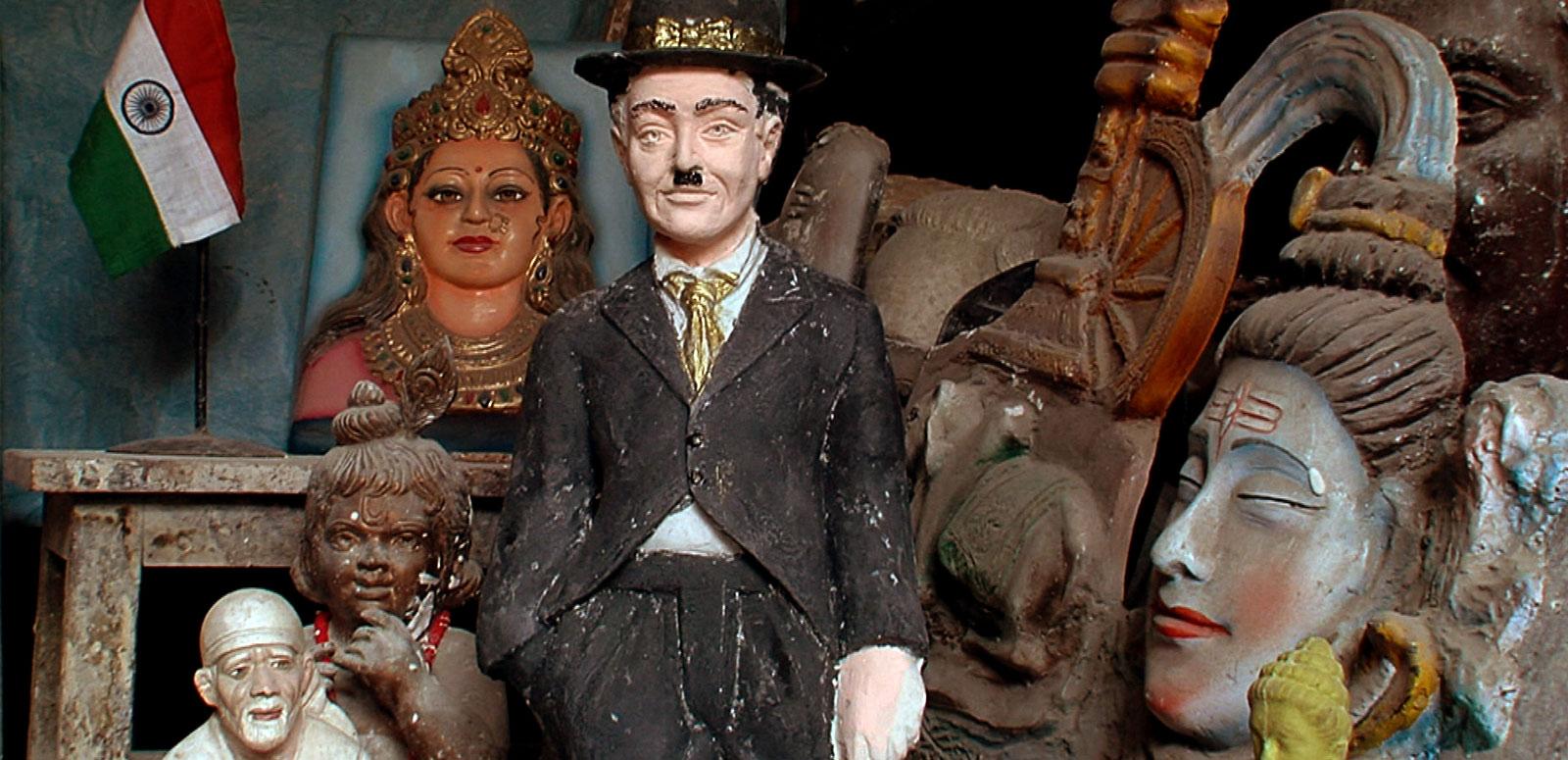 A statuette of silent film star Charlie Chaplin sitting on a shelf net to some Indian statues and trinkets and an Indian flag.