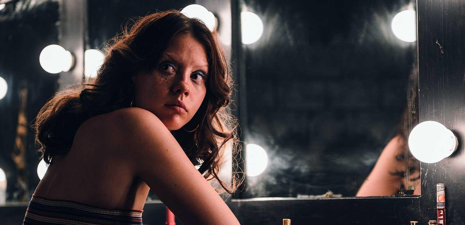 A young woman seated in front of a mirror in a dressing room. She is looking back over her right shoulder at something. 