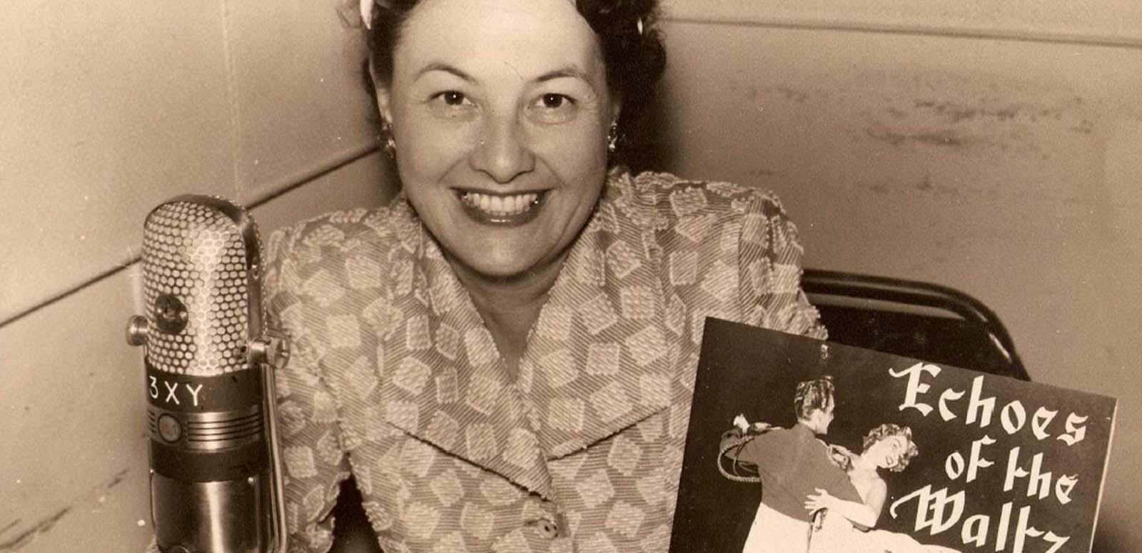 Radio personality Binny Lum behind the microphone at Radio 3XY in the 1950s or 1960s. She holds a vinyl record labelled Echoes of the Waltz.