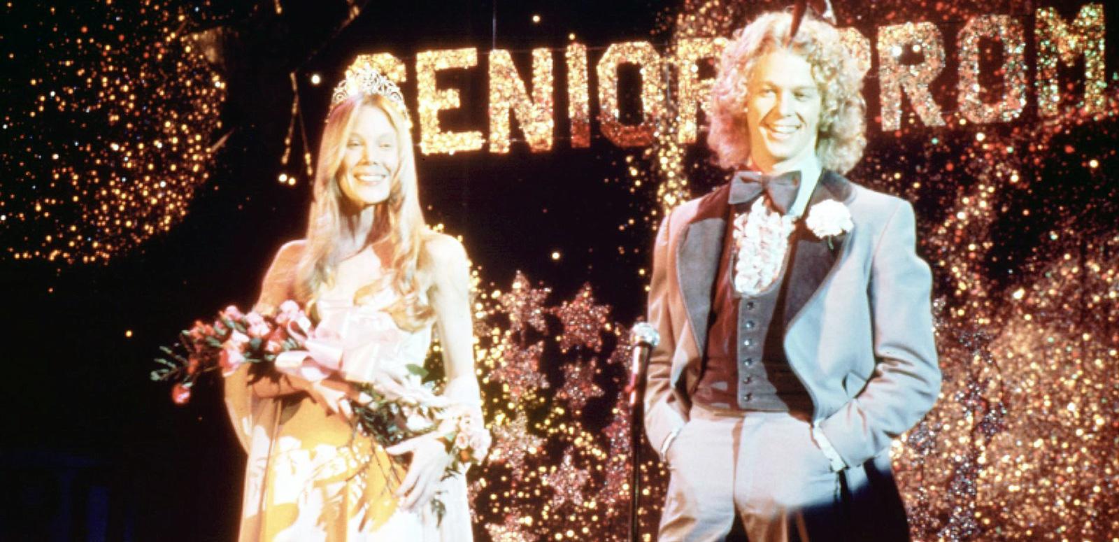A teenage girl and boy at Senior Prom. The girl is wearing a tiara and holding a bunch of flowers. The boy is wearing a tuxedo. They are both smiling.