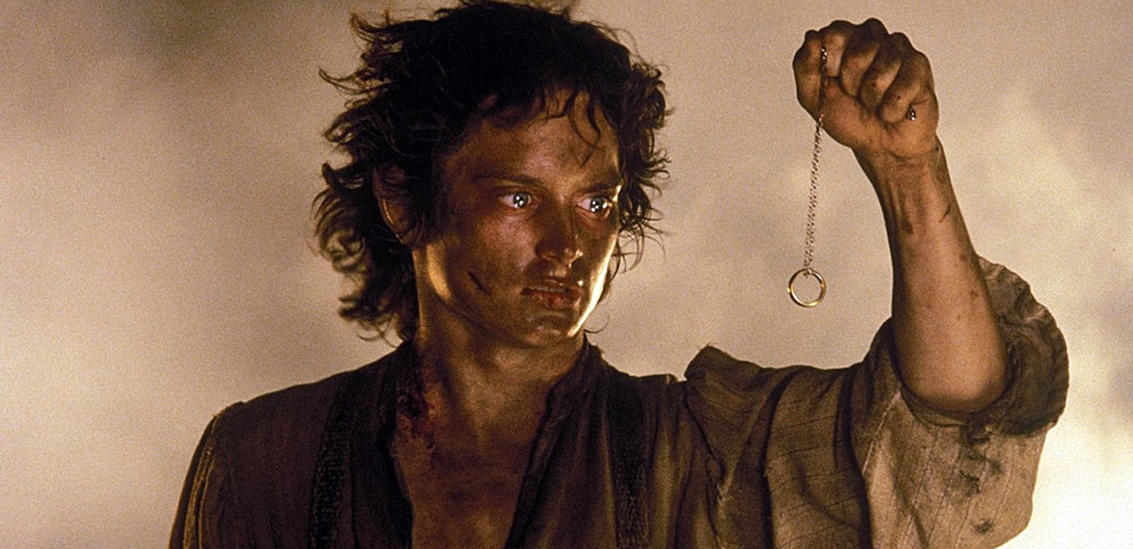 A scene from The Lord of the Rings: The Return of the King showing a head and shoulders shot of Frodo (Elijah Wood) holding up a chain with a ring dangling from it. He is covered in dirt and there is thick smoke behind him.