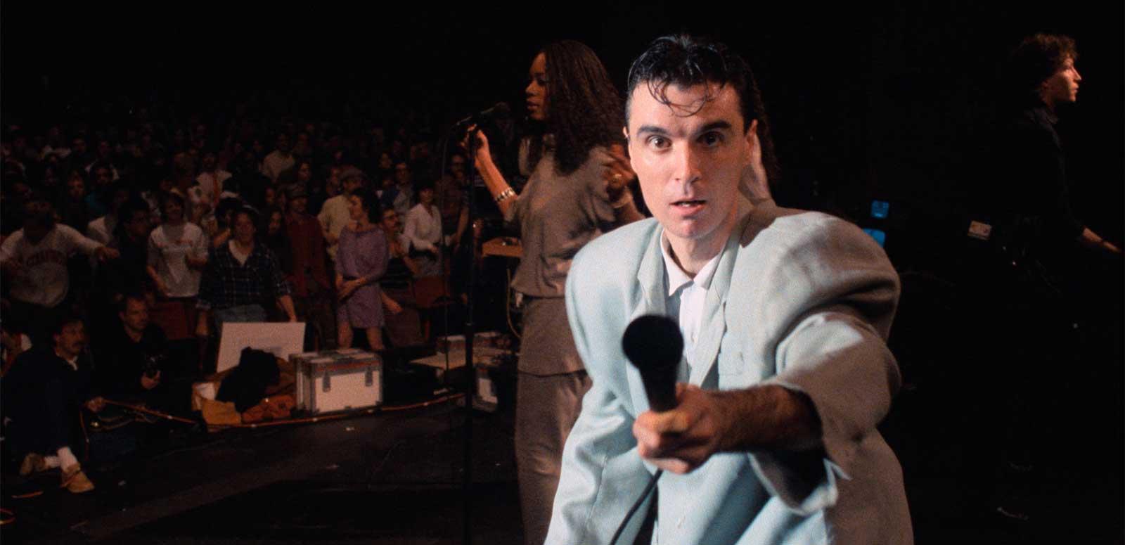 Singer David Byrne on stage with an audience behind him.  He is looking at the camera and holding out a microphone towards it.