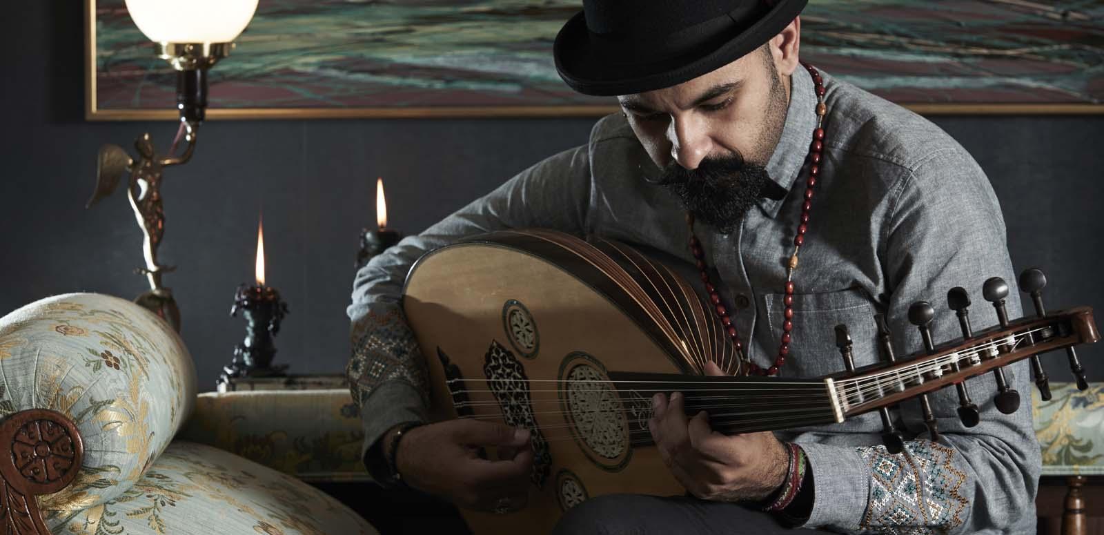 Joseph Tawadros wearing a black bowler hat and seated on a divan, playing an oud
