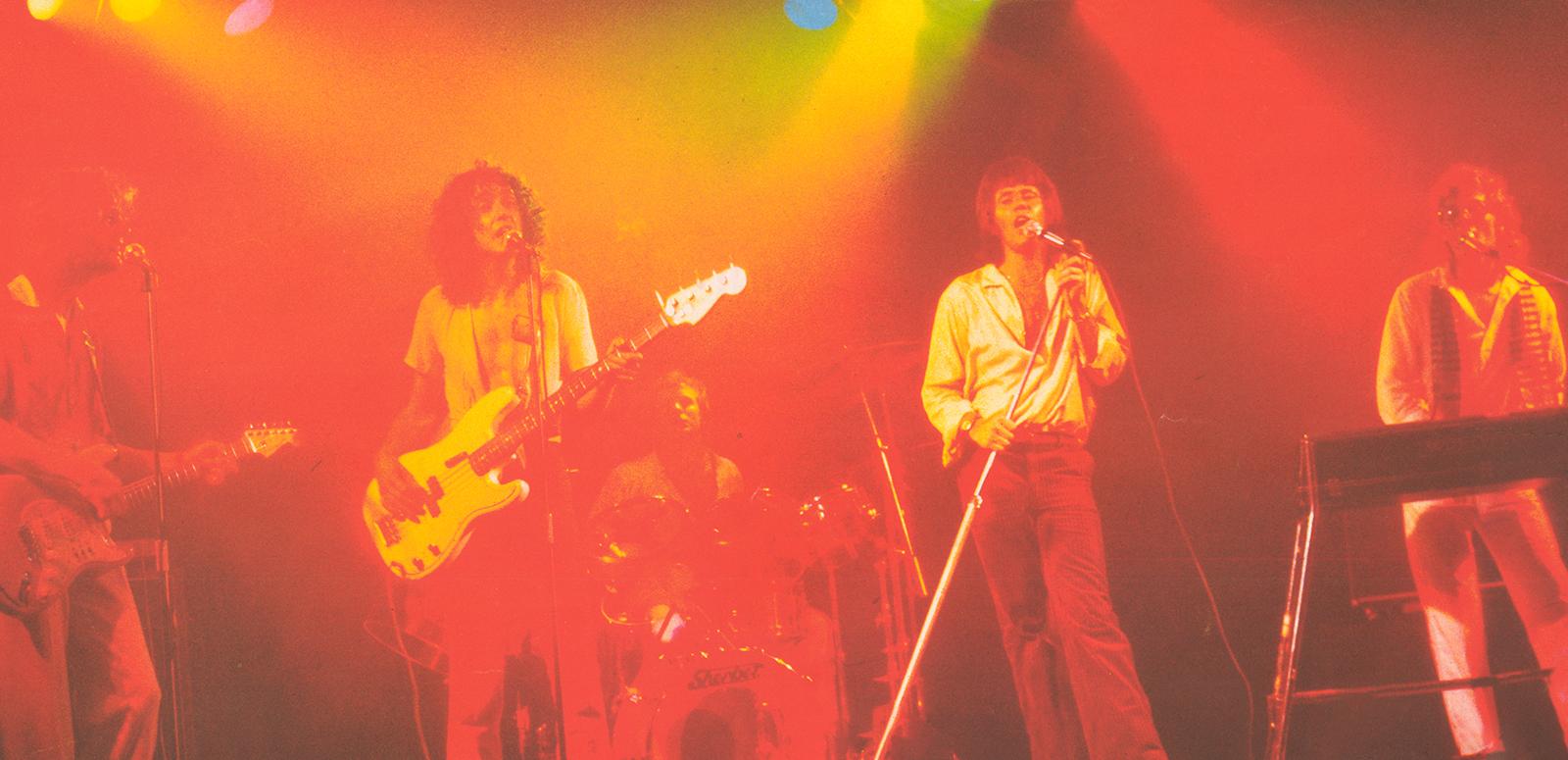 The 5 male members of 1970s rock band Sherbet performing on stage: a lead guitarist, bass guitarist, drummer and keyboards player with lead singer Daryl Braithwaite
