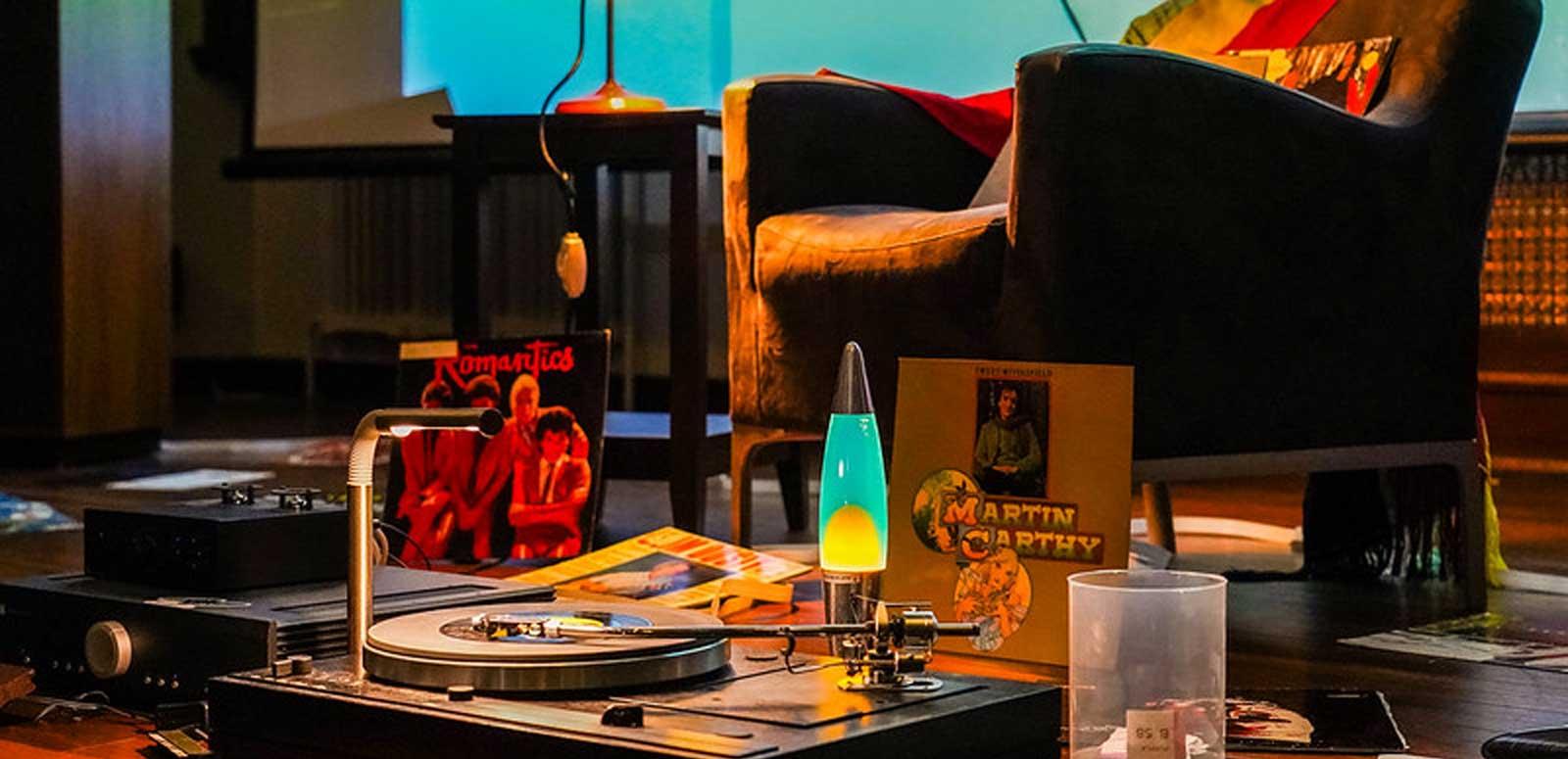 A table that has a turntable and records on it and a lava lamp. There is an armchair in the background. 